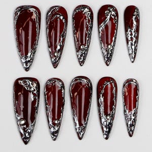 May include: Set of ten burgundy-colored, almond-shaped press-on nails. Each nail features a silver, textured border and a small, silver accent. The nails have a glossy finish and are arranged on a white surface.