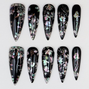 Puede incluir: Juego de diez uñas postizas negras con diseños florales iridiscentes. Cada uña tiene un acabado brillante y un patrón floral diferente en tonos rosa, verde y plateado. Las uñas tienen forma de almendra.