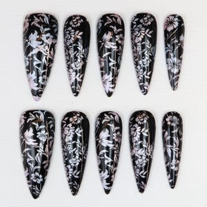 Könnte beinhalten: Ein Set aus zehn schwarzen Kunstnägeln mit glänzendem Finish. Jeder Nagel ist mit einem zarten weißen Blumenmuster verziert. Die Nägel variieren in Größe und Form, mit einem spitz zulaufenden Mandeldesign.
