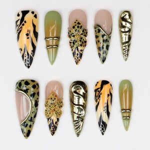 May include: A collection of ten long, almond-shaped artificial nails. Designs include animal print, floral accents, and metallic gold embellishments. Colors include black, yellow, green, and nude. The nails are arranged on a white surface.