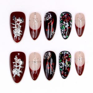 May include: A set of ten artificial nails in various designs. Some nails are burgundy with silver floral accents, others have a nude base with a burgundy tip and silver starburst, and some feature a rose design.