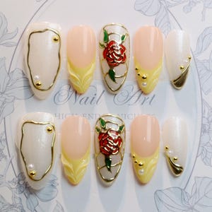 May include: A set of ten artificial nails with various designs. Some nails feature a pearlescent white base with gold accents, while others have a pale pink base with yellow and gold detailing. One nail design includes a red rose.