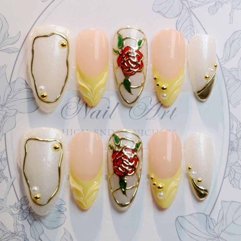 May include: A set of ten artificial nails with various designs. Some nails feature a pearlescent white base with gold accents, while others have a pale pink base with yellow and gold detailing. One nail design includes a red rose.