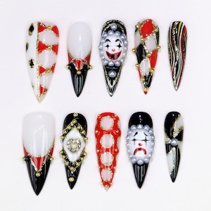 May include: A set of ten long, almond-shaped artificial nails. Designs include harlequin patterns, clown faces, and abstract gold and silver accents. The color palette features red, black, white, and gold, with pearl embellishments.