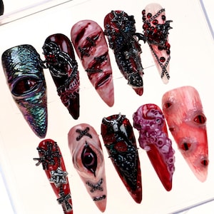 Puede incluir: Una colección de uñas postizas largas y puntiagudas con temática gótica. Las uñas presentan diseños intrincados, incluyendo ojos, cruces y otros elementos oscuros. Los colores incluyen rojo, negro y verde iridiscente.
