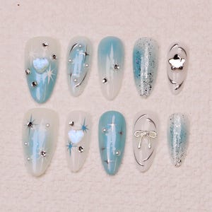 Può includere: Dieci unghie finte ombré blu chiaro e bianco con glitter argentati, strass e ciondoli argentati. I ciondoli includono un cuore, una stella e un fiocco.