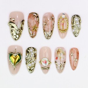 May include: A collection of ten almond-shaped artificial nails. Each nail features a nude base with gold embellishments, including swirls, stars, pearls, and miniature cherub artwork. Some nails have iridescent heart and gem accents.