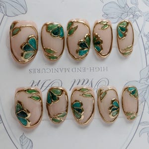 Pu&ograve; includere: Set di dieci unghie finte con base rosa chiaro e disegni floreali dorati e verdi. Le unghie sono di forma ovale e hanno una finitura lucida. Il testo "HIGH-END MANICURES" &egrave; stampato sullo sfondo.