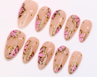 Puede incluir: Juego de uñas postizas en forma de almendra. Cada uña es de color nude con diseños florales rosas y detalles dorados. También hay pequeños puntos dorados en cada uña. Las uñas están dispuestas sobre una superficie blanca.