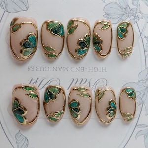 Pu&ograve; includere: Set di dieci unghie finte ovali con una base rosa chiaro e disegni floreali dorati e verdi. Le unghie sono disposte in due file su una superficie bianca con il testo "HIGH-END MANICURES" stampato sulla superficie.