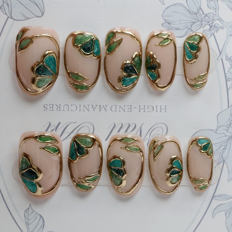 Pu&ograve; includere: Set di dieci unghie finte ovali con una base rosa chiaro e disegni floreali dorati e verdi. Le unghie sono disposte in due file su una superficie bianca con il testo "HIGH-END MANICURES" stampato sulla superficie.
