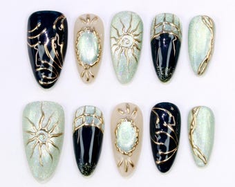 May include: A collection of ten almond-shaped false nails. The nails showcase diverse designs, such as gold sunbursts, geometric patterns, and ornate gold accents. Colours include black, beige, and iridescent green, set against a white background.