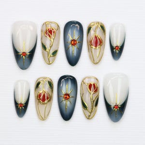May include: Ten false fingernails with a white and blue ombre design. Some nails have a gold star design, while others have a gold floral design with a red flower.
