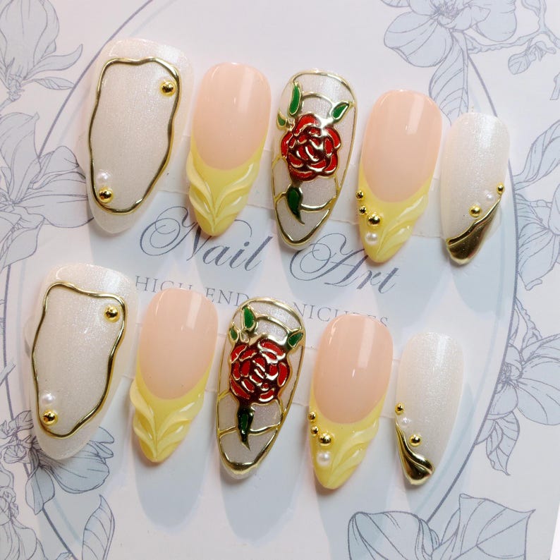 May include: A set of ten artificial nails with various designs. Some nails are white with gold accents, others have a light pink base with yellow tips. Two nails feature a red rose design with green leaves. The text "Nail Art" is visible.