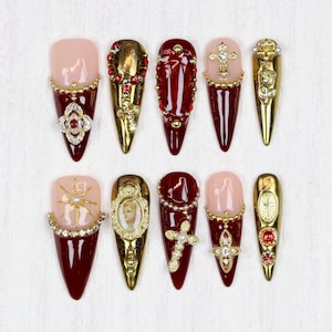 Pode incluir: Uma coleção de dez unhas artificiais longas em forma de amêndoa. Cada unha apresenta um design único com uma combinação de cores, incluindo dourado, vermelho e nude. Os enfeites incluem strass, cruzes e detalhes dourados ornamentados, criando uma estética luxuosa.