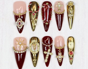 Könnte beinhalten: Eine Sammlung von zehn langen, mandelförmigen Kunstnägeln. Jeder Nagel hat ein einzigartiges Design mit einer Kombination aus Gold, Rot und Nude. Verzierungen umfassen Strasssteine, Kreuze und verzierte goldene Akzente, die eine luxuriöse Ästhetik schaffen.