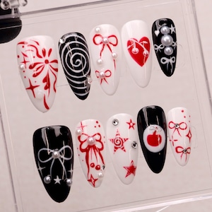Può includere: Set di dieci unghie finte con vari design. Le unghie sono bianche e nere con accenti rossi. Alcuni design includono un vortice, una stella, un fiocco e un cuore. Le unghie sono decorate con piccole perle bianche.
