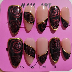 May include: A set of press-on nails with a rose design. The nails feature a black base with a red rose pattern, accented with gold trim. Some nails are nude with a black and red rose design and gold trim. The text "NAIL ART" is visible.