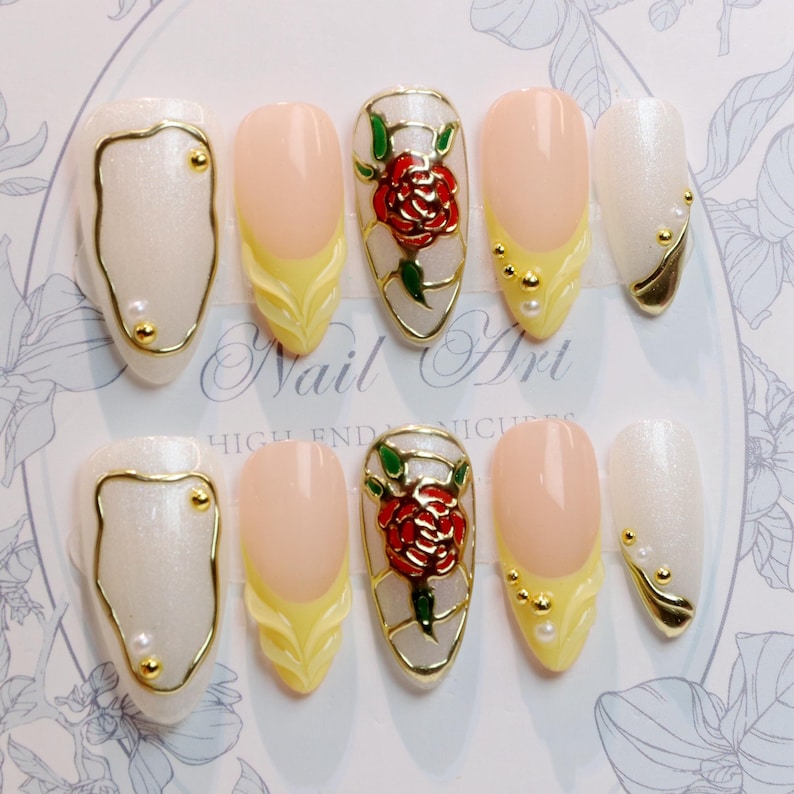 May include: A set of ten artificial nails with various designs. The nails feature a pearlescent white, pale pink, and yellow color scheme, accented with gold trim and embellishments. Some nails showcase a red rose design.