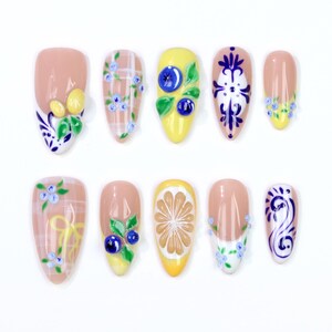 May include: A set of ten artificial nails with various designs. The nails feature a nude base with detailed artwork, including floral patterns, fruit slices, and decorative swirls. Colors include yellow, blue, green, and white.