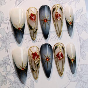 May include: A set of ten long, almond-shaped artificial nails. The nails feature a gradient design from white to dark blue, with gold accents and red floral designs. Each nail has a gold starburst design with a red gem.