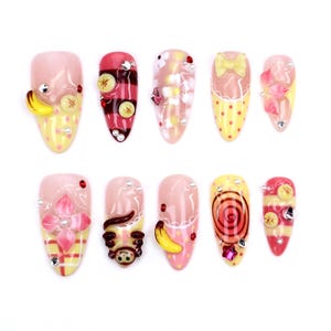 Könnte beinhalten: Ein Set aus zehn künstlichen Nägeln mit verschiedenen Designs. Die Nägel weisen eine Reihe von Farben auf, darunter Rosa, Gelb und Rot, und sind mit Verzierungen wie Früchten, Schleifen und Strasssteinen versehen. Die Nägel sind für Nail Art konzipiert.