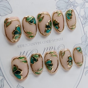 Pu&ograve; includere: Set di dieci unghie finte ovali con una base rosa chiaro e disegni floreali dorati e verdi. Le unghie sono disposte in due file su uno sfondo bianco con il testo "HIGH-END MANICURES" stampato sullo sfondo.