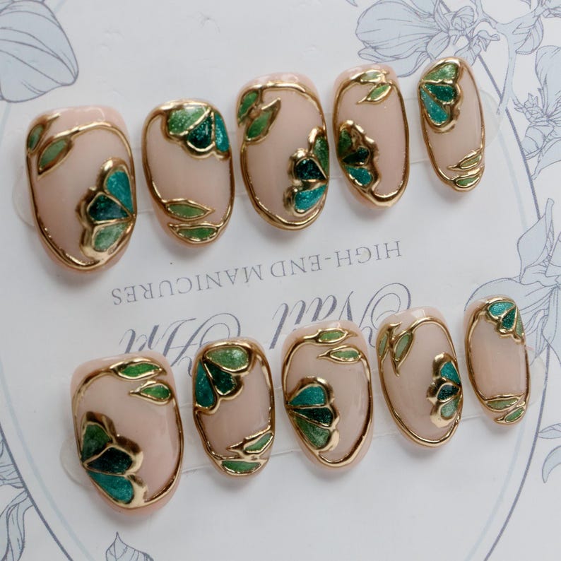 Pu&ograve; includere: Set di dieci unghie finte ovali con una base rosa chiaro e disegni floreali dorati e verdi. Le unghie sono disposte in due file su uno sfondo bianco con il testo "HIGH-END MANICURES" stampato sullo sfondo.