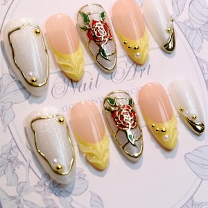 Könnte beinhalten: Ein Set künstlicher Nägel mit verschiedenen Designs. Einige Nägel sind perlmuttweiß mit goldenen Verzierungen und Akzenten. Andere haben ein gelb-pfirsichfarbenes Design, und einige haben ein rotes Rosendesign mit goldenen Verzierungen. Der Text "Nail Art" ist sichtbar.