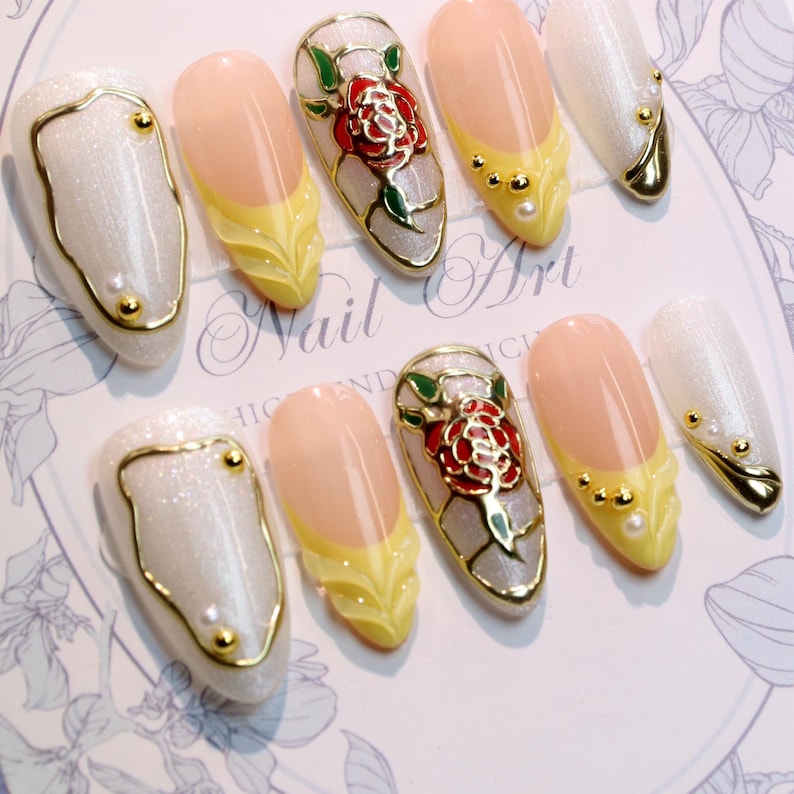 May include: A set of artificial nails with various designs. Some nails are pearlescent white with gold trim and accents. Others feature a yellow and peach design, and some have a red rose design with gold trim. The words "Nail Art" are visible.