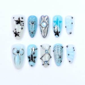 Peut inclure: Un ensemble de dix faux ongles aux designs variés. Les ongles présentent un mélange de couleurs blanc et bleu clair, ornés de nœuds argentés, d'étoiles et de motifs géométriques. Certains ongles ont un motif tourbillon ou de petits ornements sombres.