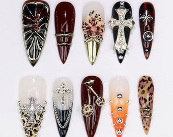 May include: A collection of ten long, pointed artificial nails. Designs include crosses, leopard print, cherries, and floral accents. Colors include burgundy, black, gold, and clear. Some nails feature rhinestones and metallic embellishments.