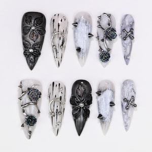 May include: A set of ten long, pointed artificial nails. Designs include black floral accents, silver spikes, and silver metal embellishments. The nails feature a mix of black, white, and gray color schemes, with a glossy finish.
