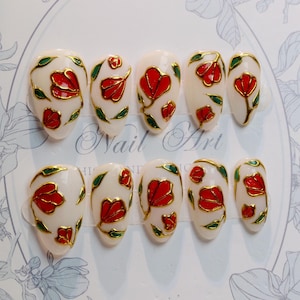 May include: A set of ten artificial nails with a cream-colored base. Each nail features a red flower design with gold outlines and green leaves. The words "Nail Art" are visible in the background.