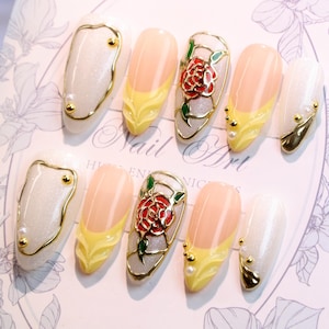 May include: A set of artificial nails with various designs. Some nails feature a gold outline with pearl accents, others have a yellow and peach design, and some have a red rose design with green leaves. The text "Nail Art" is visible.