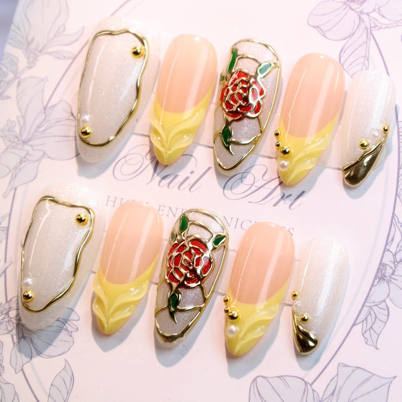 May include: A set of artificial nails with various designs. Some nails feature a gold outline with pearl accents, others have a yellow and peach design, and some have a red rose design with green leaves. The text "Nail Art" is visible.