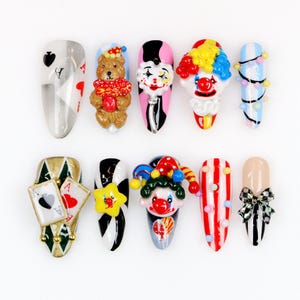 May include: A collection of ten artificial nails with various designs. The designs include playing card suits, a teddy bear, a clown, a star, and a bow. The nails feature a variety of colors, including red, white, black, blue, and gold. The nails are arranged on a white surface.