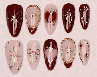 May include: Set of ten false nails with different designs. The designs include a heart, a star, and a bow. The nails are a burgundy color with gold accents.