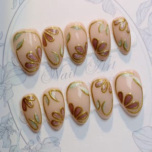 May include: A set of ten almond-shaped artificial nails. Each nail is painted with a nude base, gold outlines, and floral designs in pink and silver glitter. The words "Nail Art" are visible in the background.