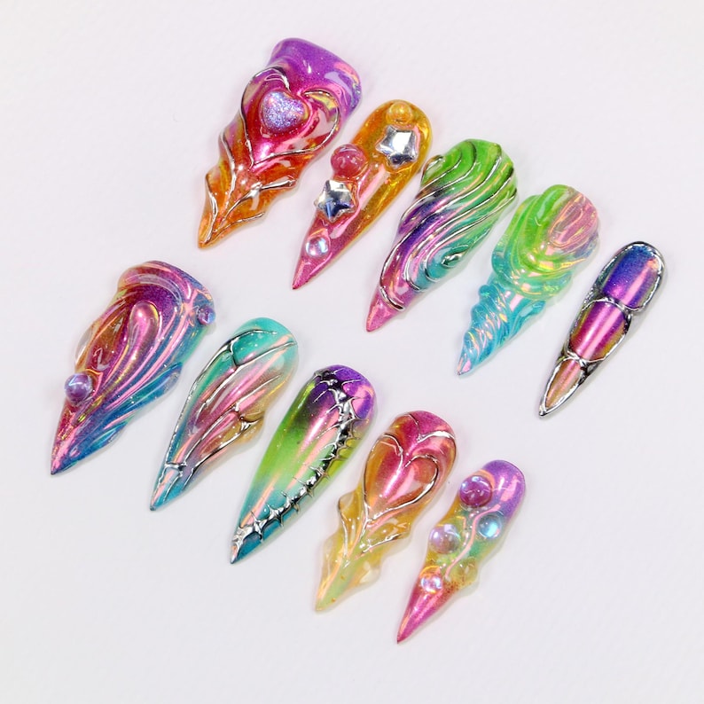 May include: A collection of ten iridescent, sculpted nail art designs. Each nail features a unique design with a variety of colors, including pink, purple, green, and gold. Some nails have embedded gems and metallic accents.