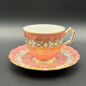 May include: A pink porcelain teacup and matching saucer set featuring an intricate gold filigree pattern. The teacup has a white interior, a pedestal base, and a gold-trimmed handle. The saucer has a scalloped edge with gold detailing.
