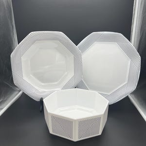May include: A set of three white octagonal ceramic dishes featuring a black linear pattern along the edges. The collection includes two plates and one deep bowl, all displaying a consistent geometric design with thin parallel black lines.