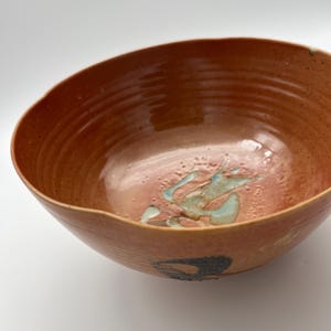 May include: A round ceramic bowl with a terracotta-coloured glaze and visible horizontal ridges along the interior walls. The base of the bowl features a textured, light blue abstract design, and a dark circular mark is visible on the exterior side.