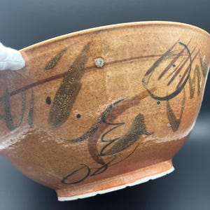 May include: A ceramic bowl with a warm, tan-coloured glaze featuring dark brown, hand-painted abstract brushstrokes and linear markings. The bowl has a rounded shape, a visible footed base, and a smooth, glossy finish across its exterior surface.