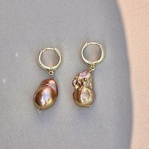 May include: A pair of gold hoop earrings featuring dangling, irregular-shaped baroque pearls with a metallic, iridescent pink and bronze luster. The pearls have a textured, organic surface and are suspended from simple, polished gold-tone metal hoops.