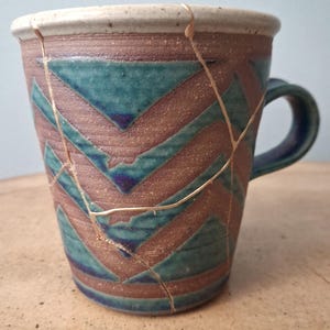 May include: A ceramic mug featuring a textured brown and teal chevron pattern with a cream-colored rim. The surface displays visible cracks repaired with gold-colored lacquer, creating a distinct network of lines across the exterior body.