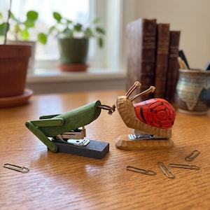 May include: Two wooden staplers shaped like animals. One is a green grasshopper with a dark gray base, and the other is a red and brown snail with a natural wood base. Both feature metal stapling mechanisms and are placed on a wooden surface.