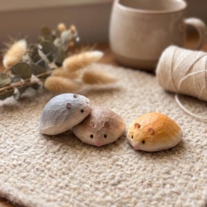 Puede incluir: Tres pequeñas figuras de animales talladas a mano en madera con forma de hámsteres redondeados. Una es gris claro, otra marrón claro y otra amarillo dorado. Cada figura presenta una textura suave y facetada, ojos oscuros y orejas pequeñas.