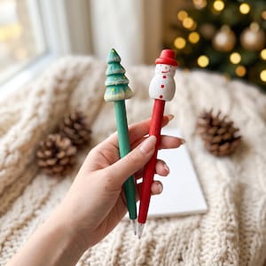 May include: Two wooden pens with festive toppers. One pen has a green body and a green Christmas tree topper with yellow dots. The other pen has a red body and a white snowman topper with a red hat, red buttons, and a painted orange nose.