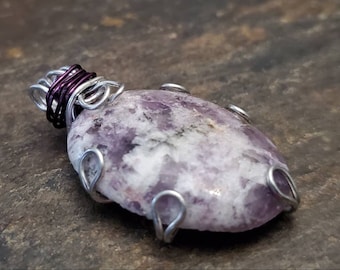 Puede incluir: Un colgante de piedra preciosa púrpura y blanca con un envoltorio de alambre plateado. El colgante tiene forma ovalada y una textura natural y rugosa.